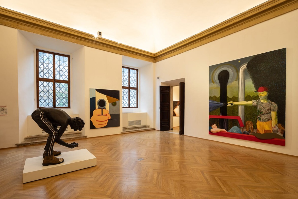 Art gallery room with a sculpture of a man bending over and two large colorful paintings on the walls.