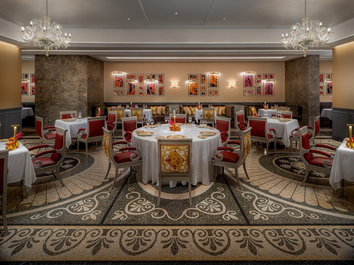 Elegant dining room with ornate chandeliers, patterned floor, and tables set with red and gold chairs and white tablecloths.