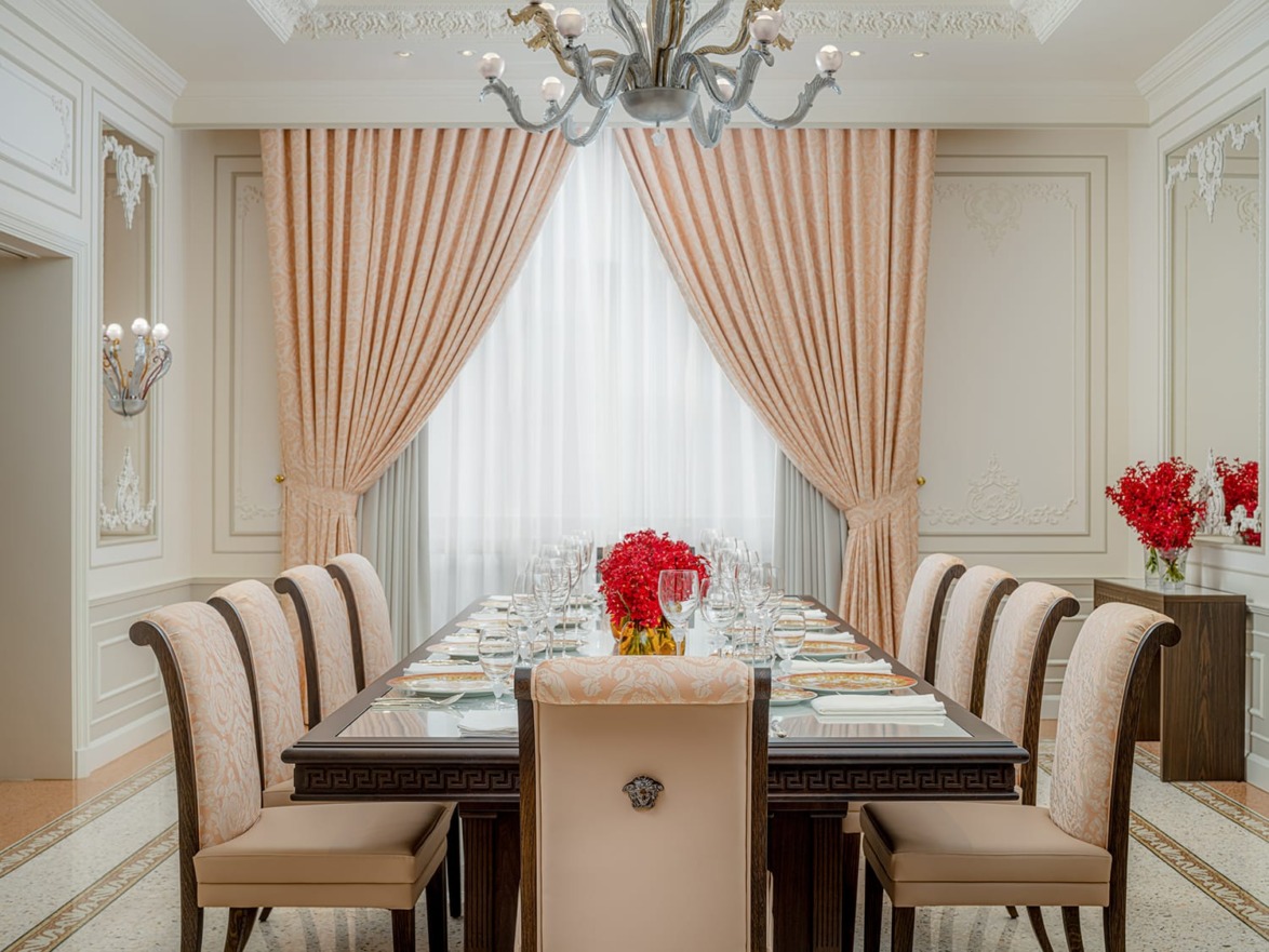 Luxurious dining room with a long table set for a meal, elegant chairs, chandeliers, and large curtains.