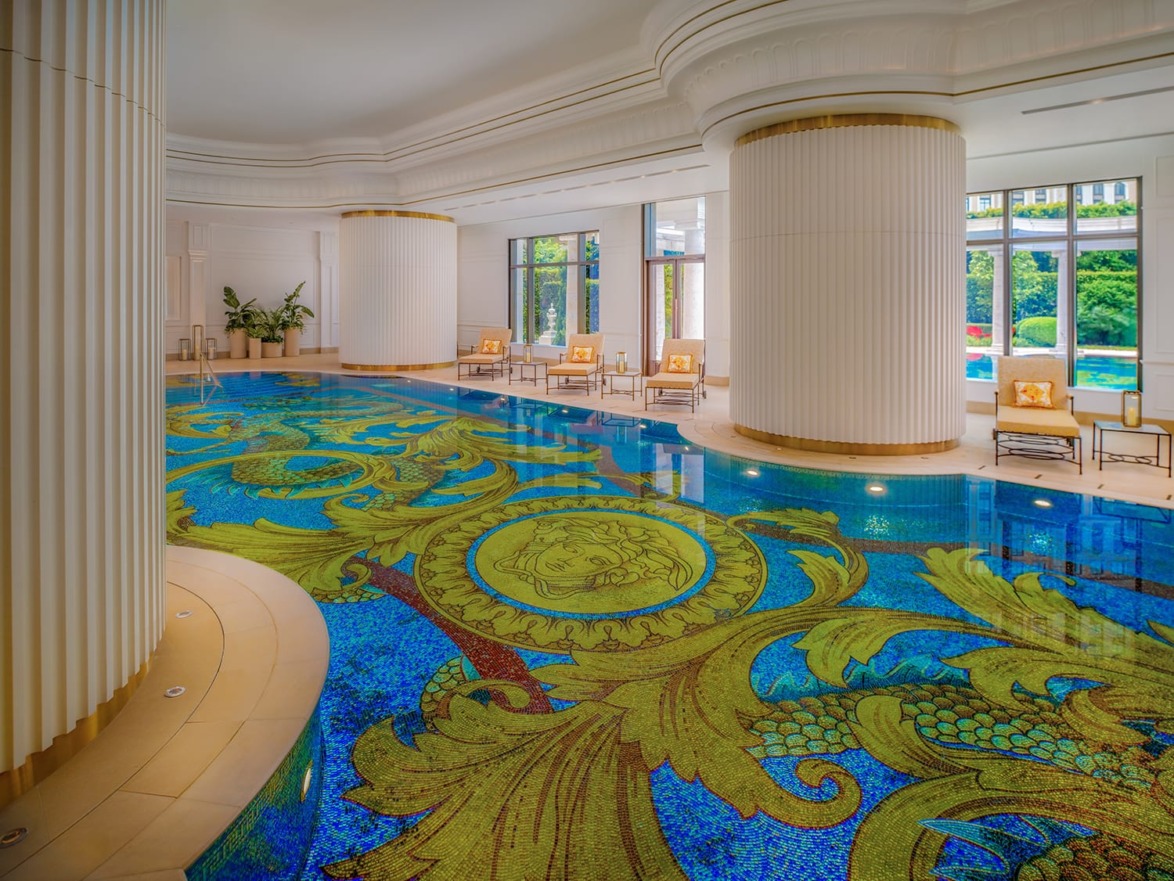 Luxurious indoor pool with ornate mosaic floor design, elegant seating, large windows, and white columns.