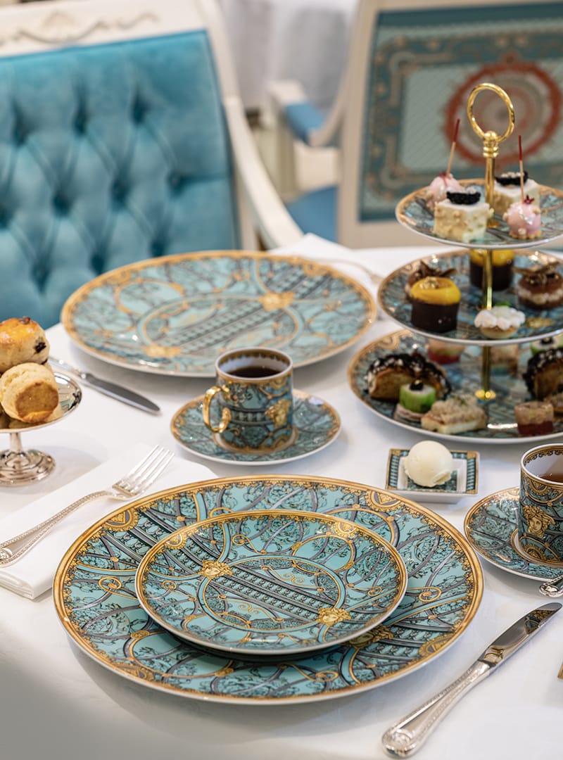 Elegant dining setup with ornate blue and gold plates, teacups, tiered tray of pastries, and a plush blue chair.