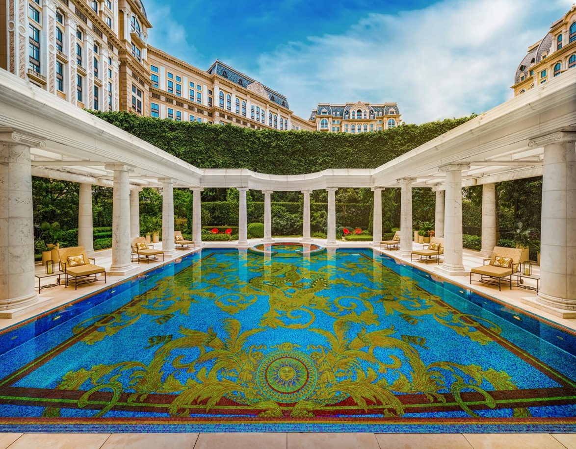 Luxurious outdoor pool with intricate mosaic designs surrounded by elegant columns and lush greenery, under a clear blue sky.