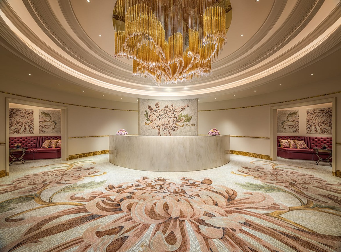 Elegant hotel lobby with large floral mosaic on the floor, curved reception desk, and ornate gold chandelier above.