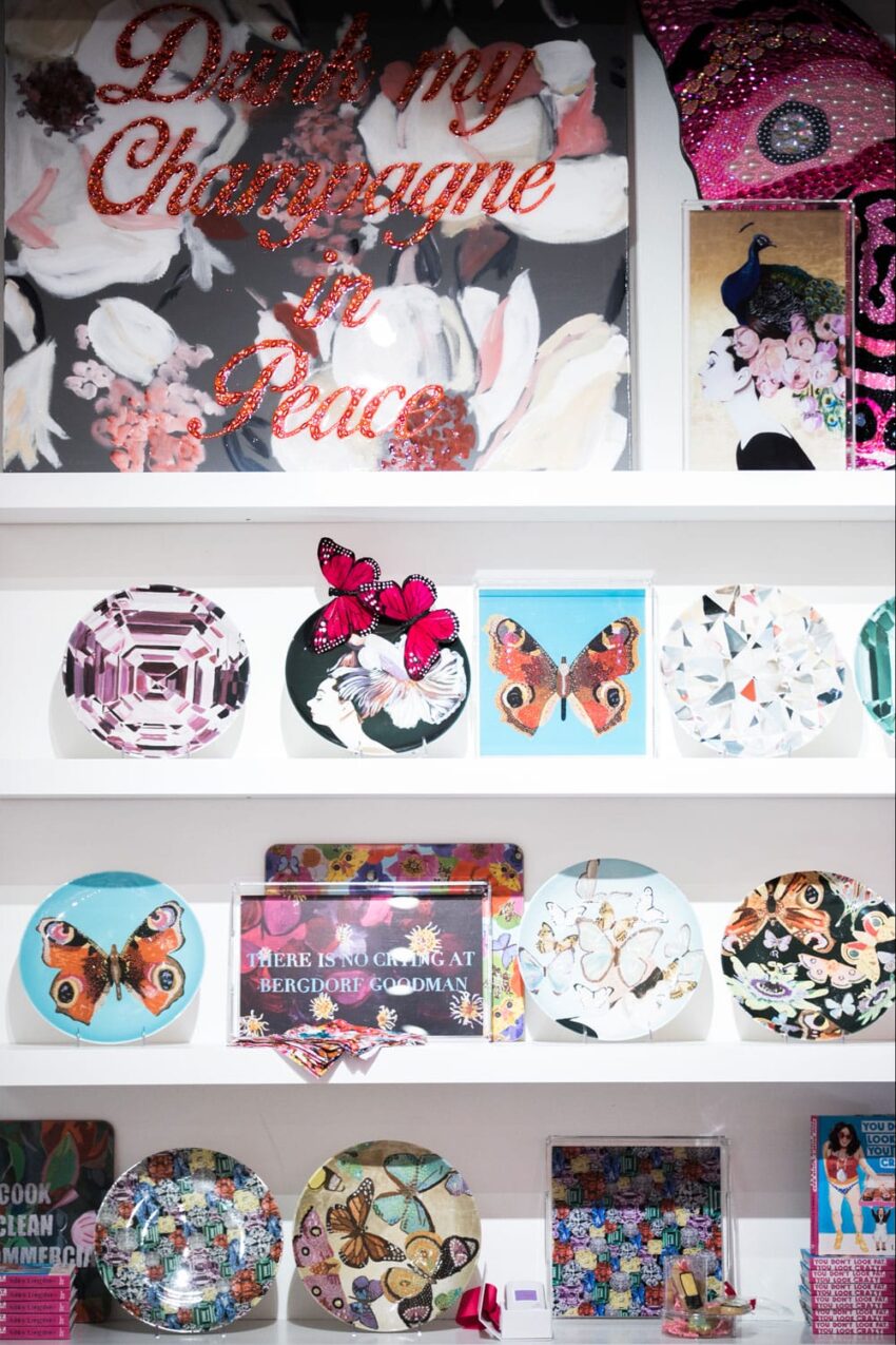 Decorative display with plates featuring butterflies and geometric patterns, topped with a "Drink my Champagne in Peace" sign.