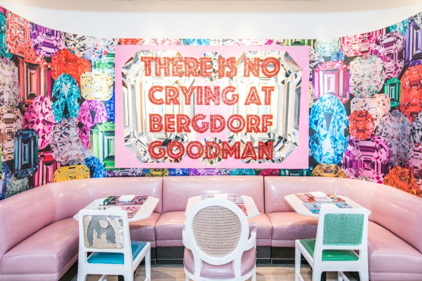 Colorful dining area with gem-themed wall art and sign reading "There Is No Crying at Bergdorf Goodman" above pink seating.