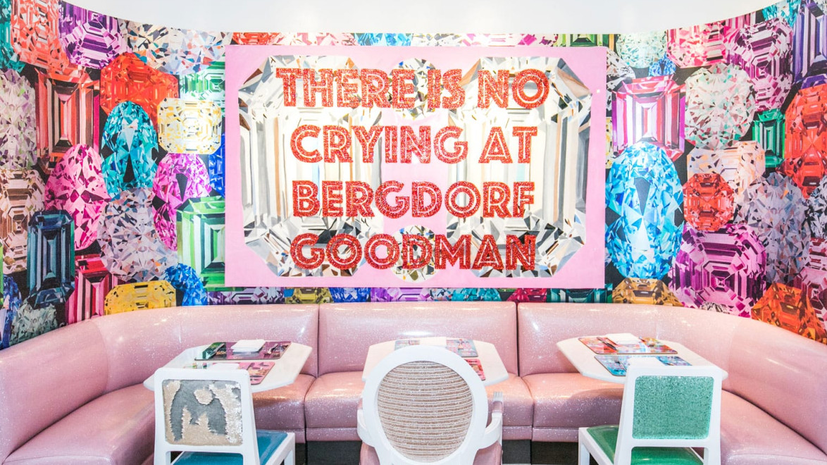 Colorful dining area with gem-themed wall art and sign reading "There Is No Crying at Bergdorf Goodman" above pink seating.