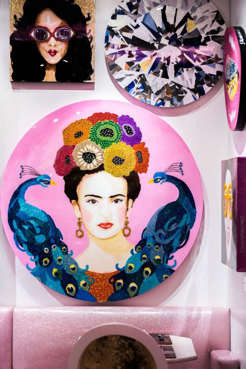 Colorful artwork featuring a woman with floral headdress, surrounded by peacocks on a pink background, displayed in a gallery.