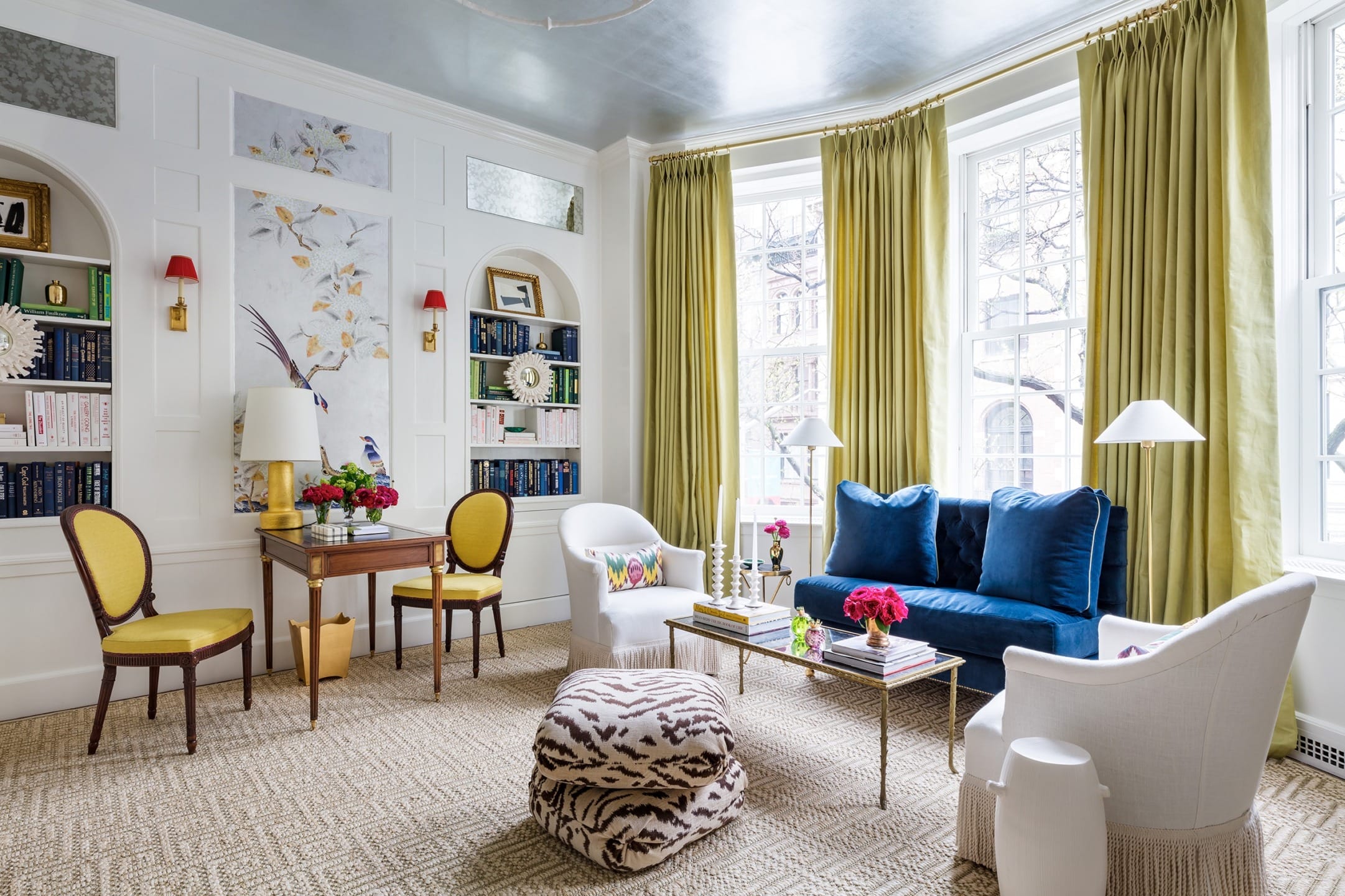 Colorful living room with yellow curtains, blue sofa, patterned ottoman, and chairs around a wooden table in natural light.
