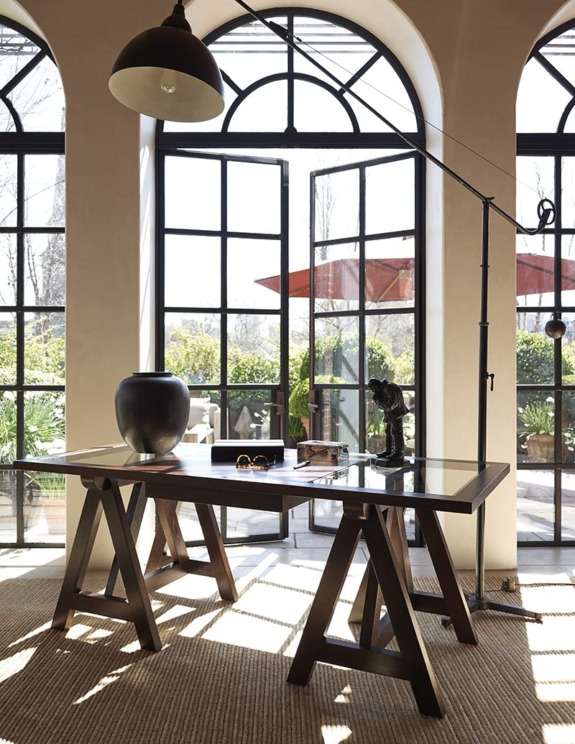 Sunlit workspace with a wooden desk, modern decor, and large arched windows overlooking a garden patio.