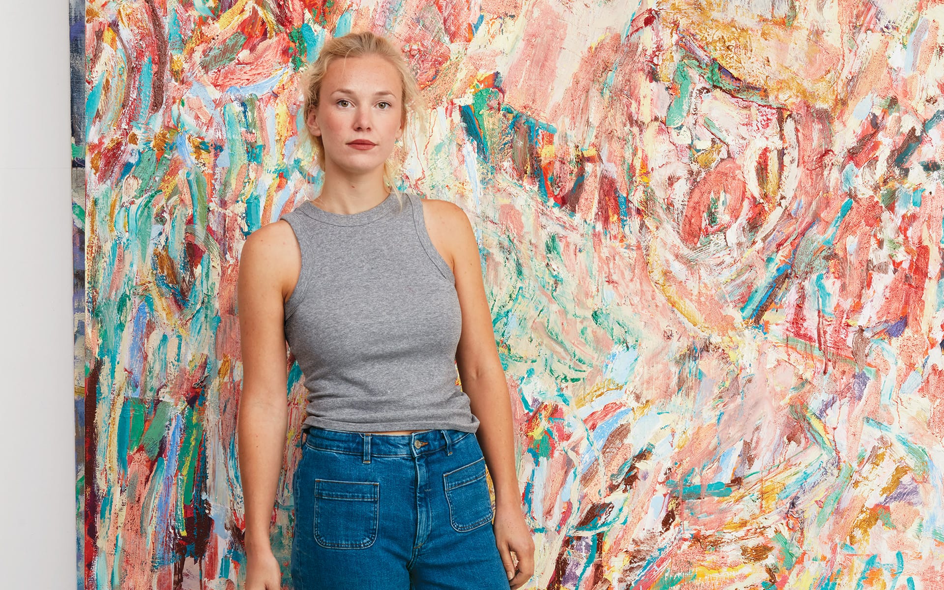 Person in a gray tank top and jeans stands in front of a colorful abstract painting with expressive brushstrokes.