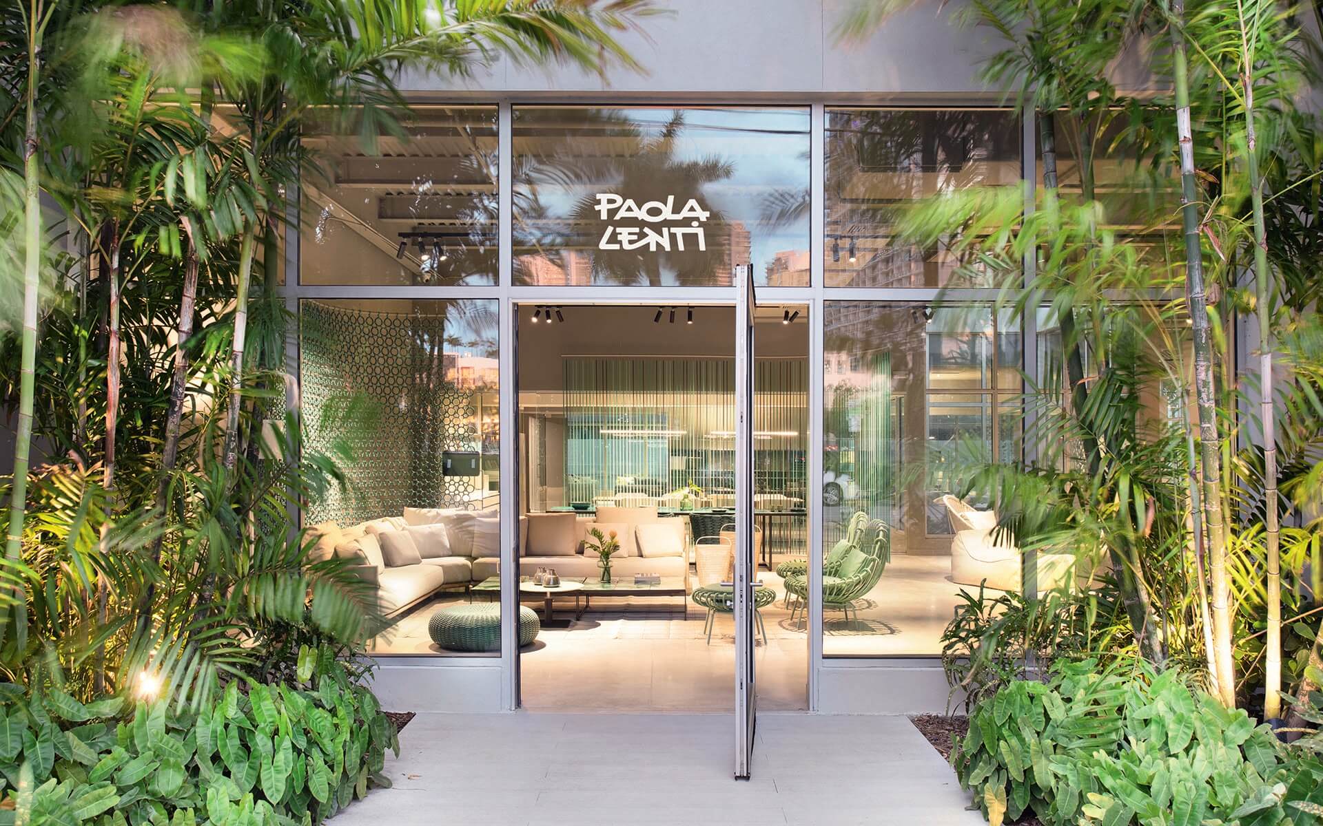Exterior view of a modern furniture store with glass front and lush green plants surrounding the entrance.