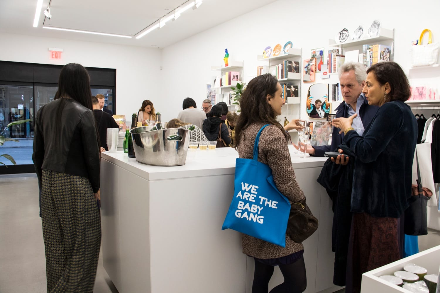People socializing in a modern store with art displays, a woman holding a blue bag reads "We Are The Baby Gang"