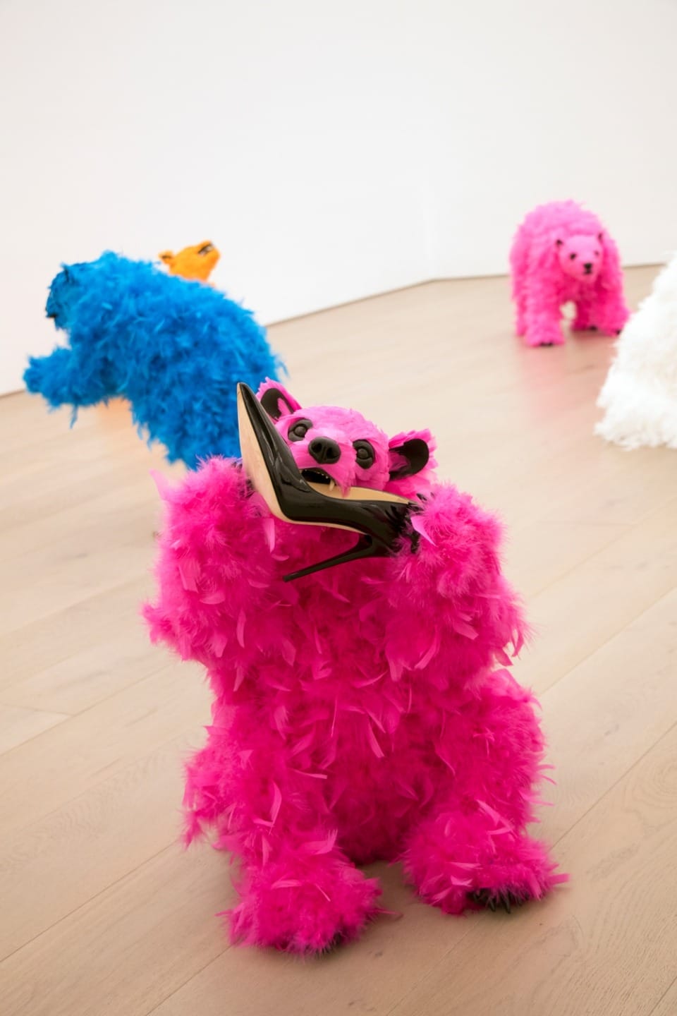 Colorful feather-covered bears posed on a wooden floor in an art exhibit, vibrant pink, blue, orange, and white fur