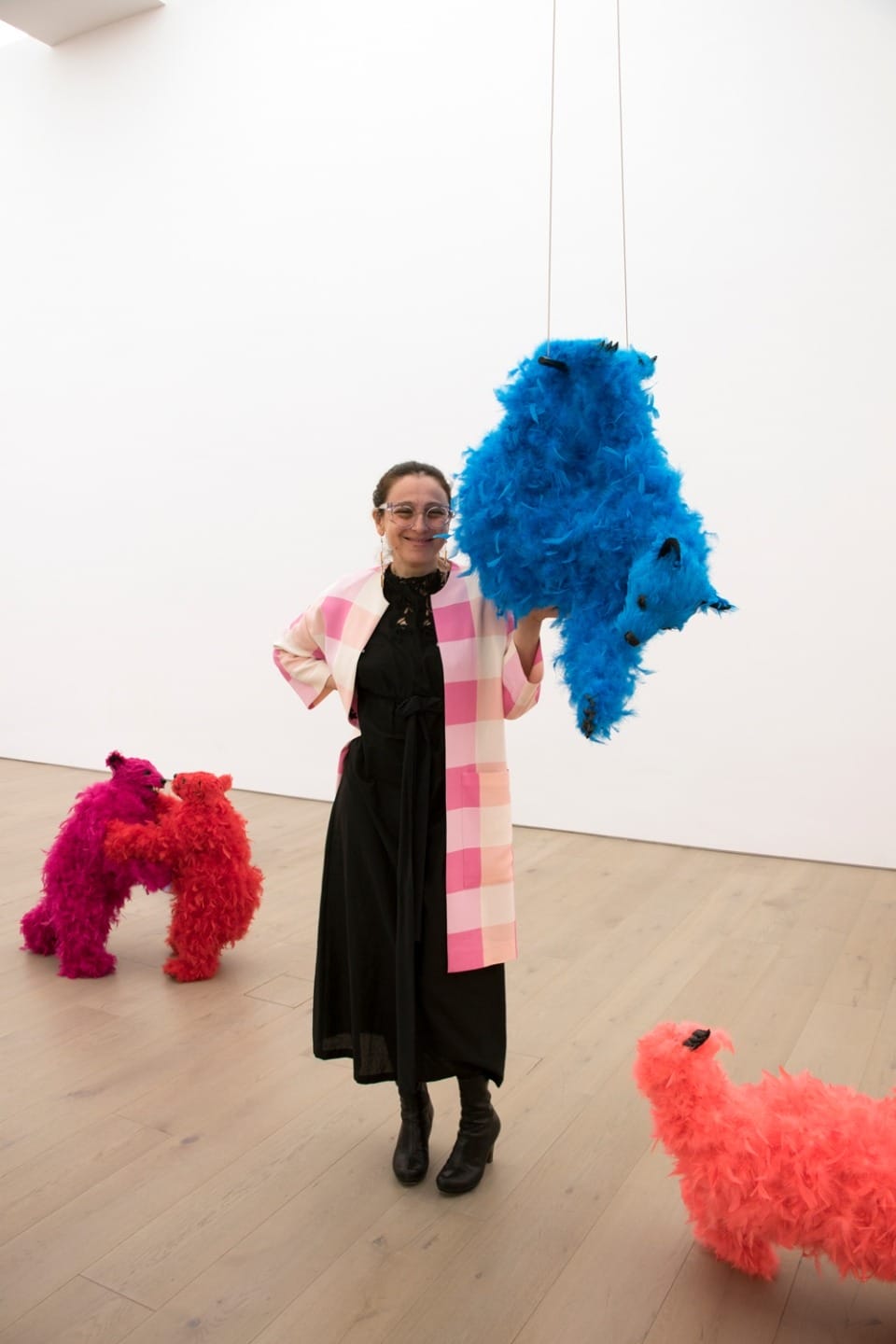 Person standing in art gallery with colorful feathered sculptures, wearing a black dress and pink plaid coat, smiling at camera.