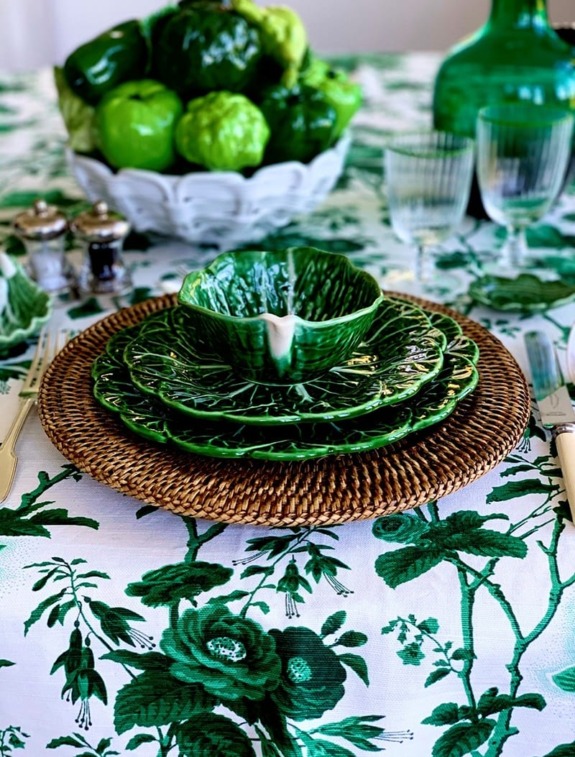 Table setting with green leaf-patterned dishes on a floral tablecloth, wicker placemat, and a centerpiece bowl of green fruits.