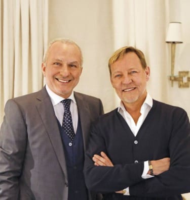 Two smiling men standing indoors, one in a suit and the other in a sweater, with neutral-colored curtains in the background.