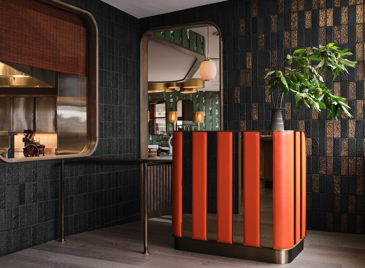 Restaurant interior with an orange reception desk, dark textured walls, and a green leafy plant in the corner.
