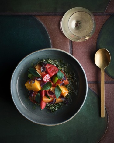 Bowl of heirloom tomato salad with basil leaves on a dark table setting next to a glass of white wine and a gold spoon.