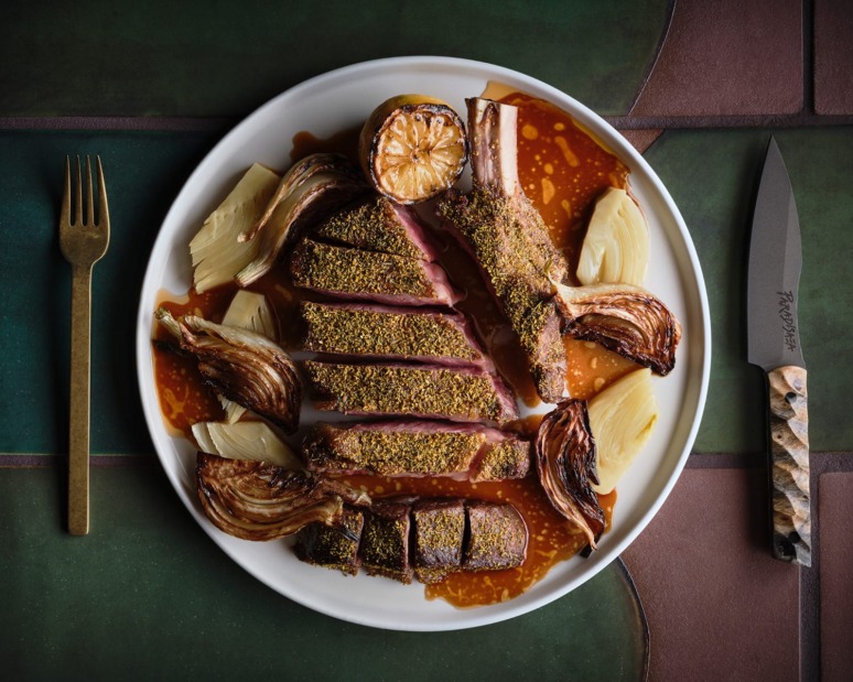 Plate of seasoned lamb chops with grilled onion, artichoke, roasted lemon, and sauce, accompanied by a knife and fork.