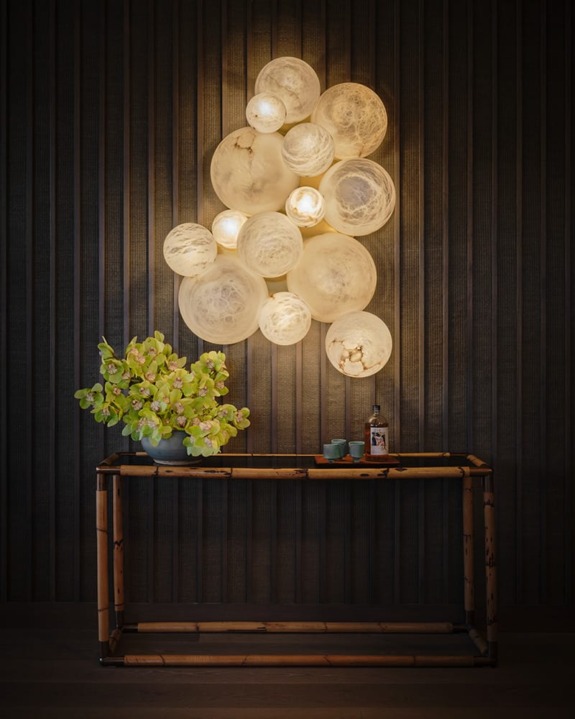 Modern wall lamp with circular bulbs above a wooden console table with a green plant and decorative items against a dark wall.