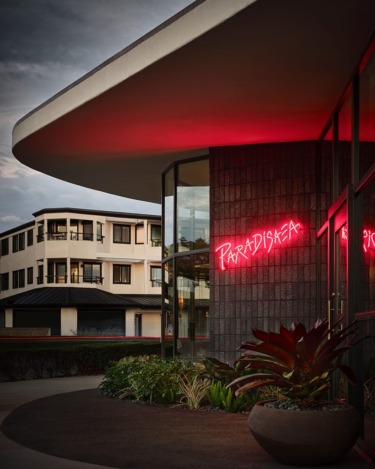 Modern building exterior with rounded roof, neon sign reading "Paradisea," and potted plants in foreground.