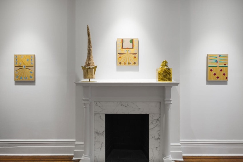 Art gallery with paintings hung on a white wall above a white marble fireplace, featuring colorful abstract designs.