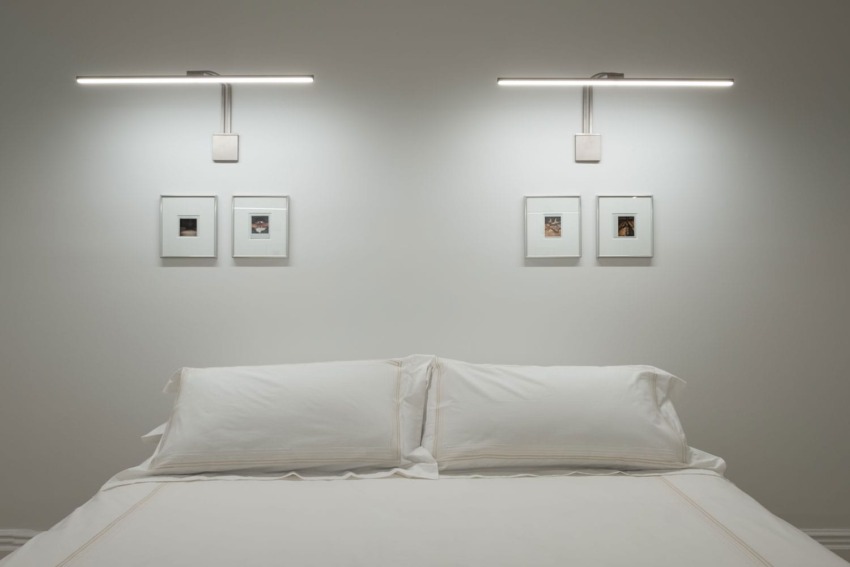 Minimalist bedroom with white bed linens, wall-mounted lights, and framed art above the headboard on a neutral-colored wall.