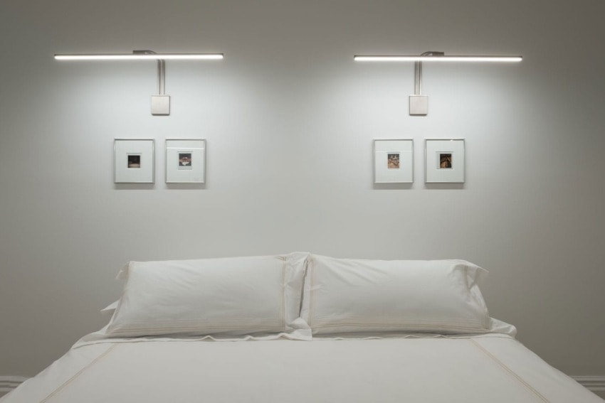 Minimalist bedroom with white bed linens, wall-mounted lights, and framed art above the headboard on a neutral-colored wall.