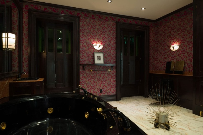 Luxurious bathroom with black bathtub, gold fixtures, red floral wallpaper, decorative lighting, and marble flooring.