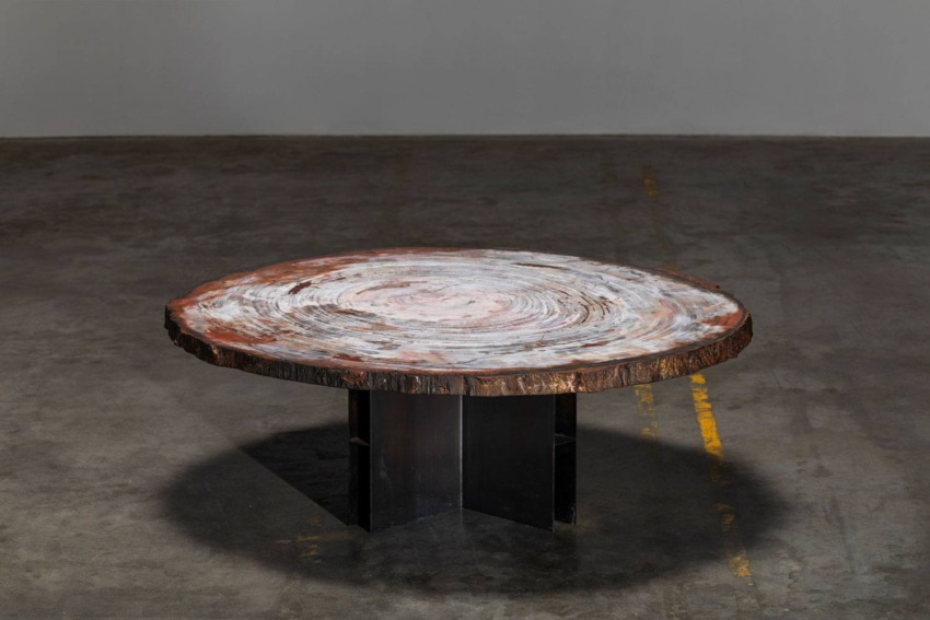 Round wooden table with a polished, textured surface on a dark pedestal base, placed on a concrete floor.