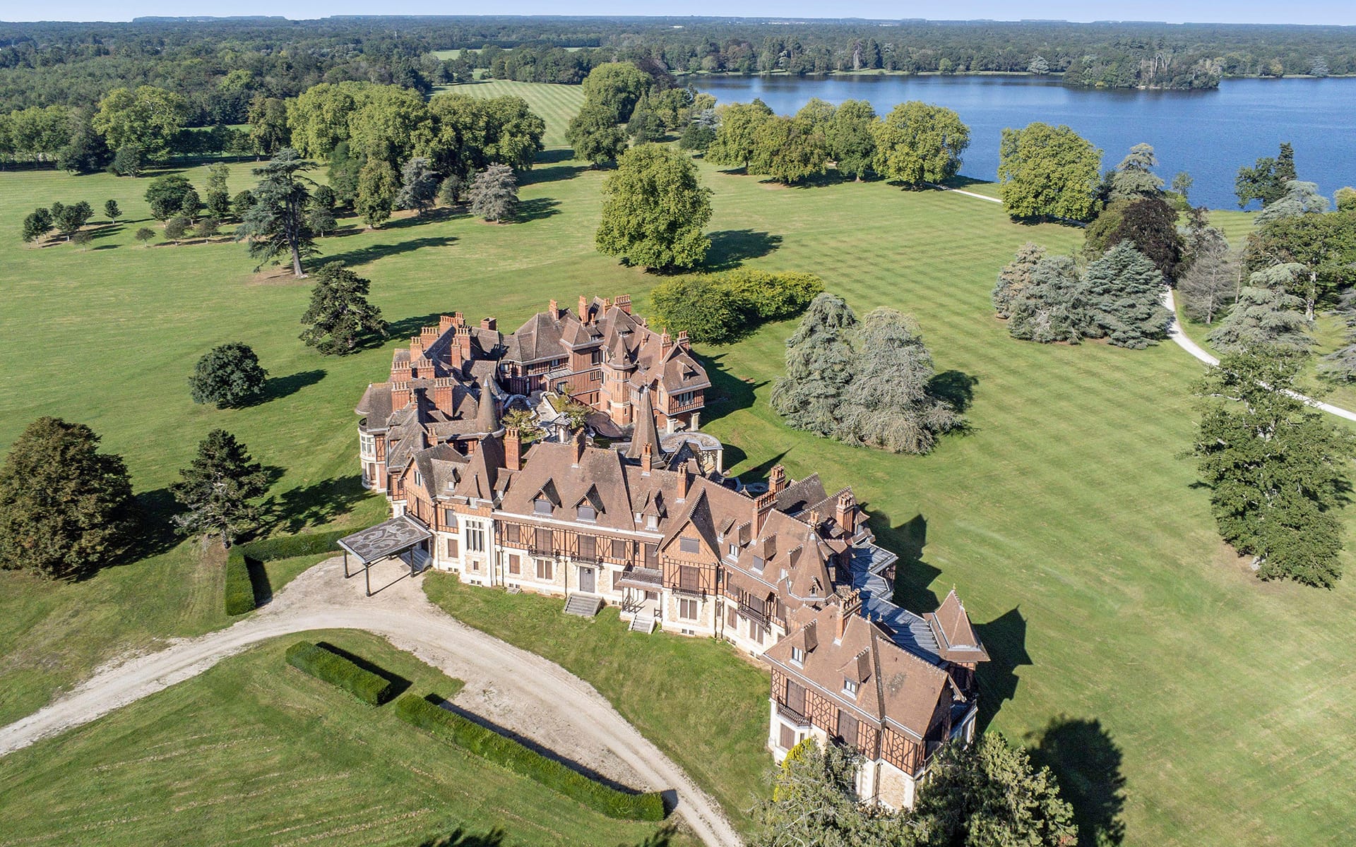 Aerial view of a large historic mansion surrounded by vast green lawns and trees near a lake.