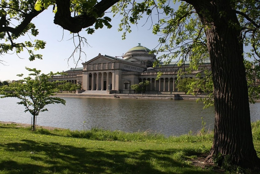 Exterior view of a classical architecture building by a lake, framed by trees and greenery on a sunny day.