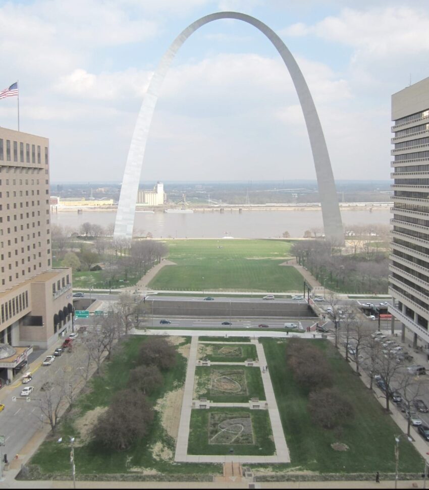 View of Gateway Arch in St. Louis with park and buildings in foreground on a partly cloudy day.