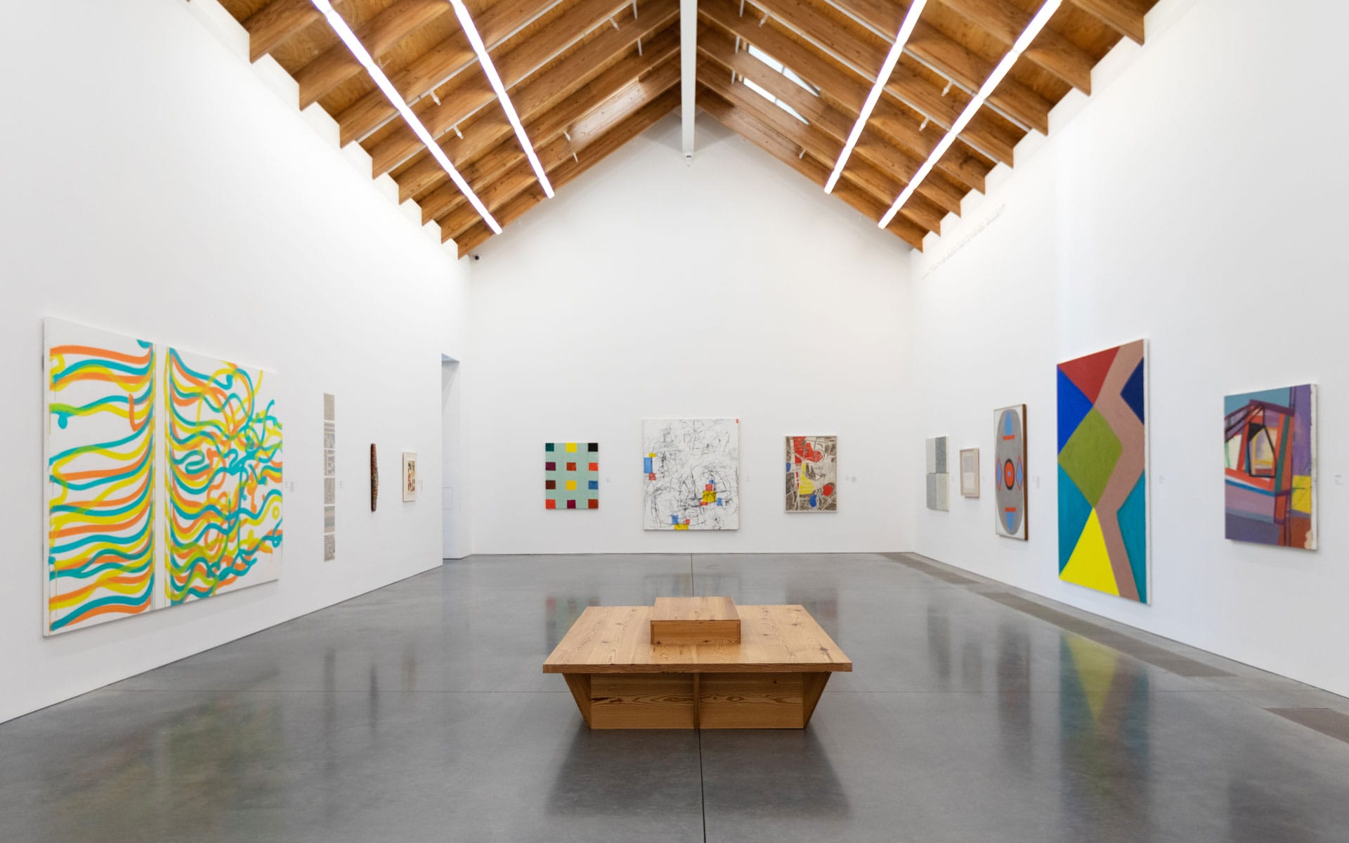 Contemporary art gallery with colorful abstract paintings on white walls and a wooden bench in the center of the room.