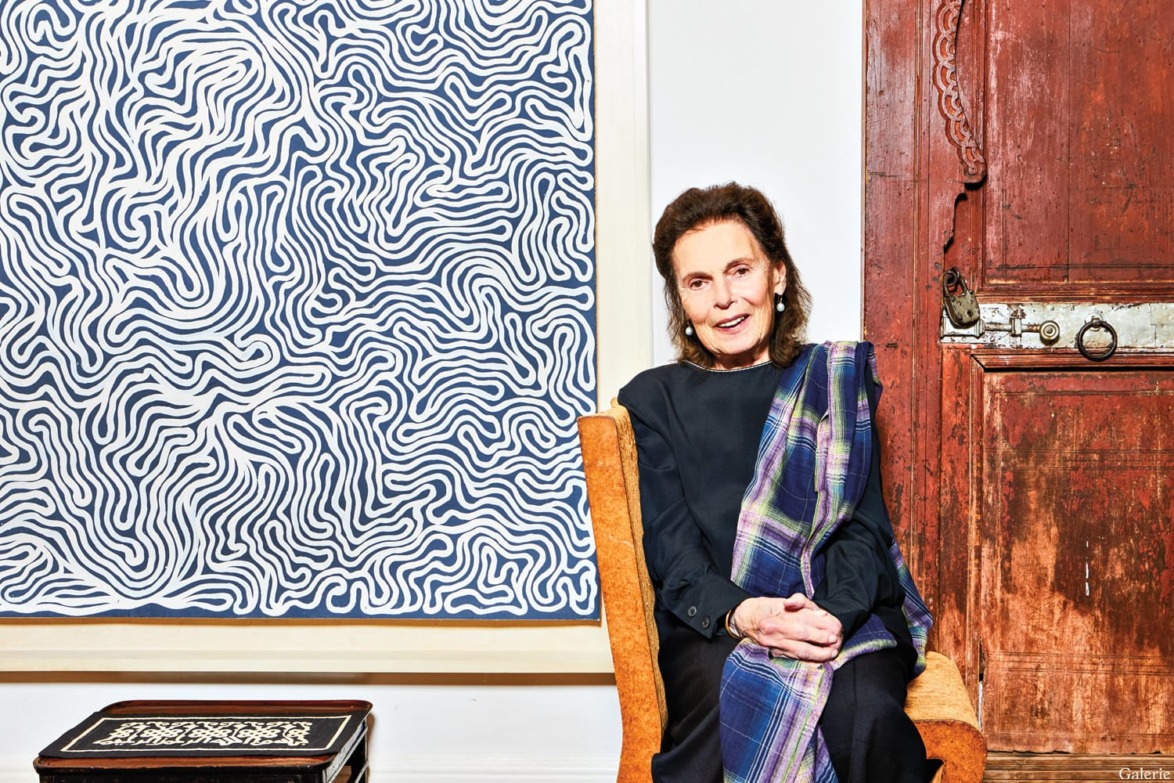 Person sitting on a chair next to abstract art and a wooden door, wearing a patterned shawl and dark clothing.