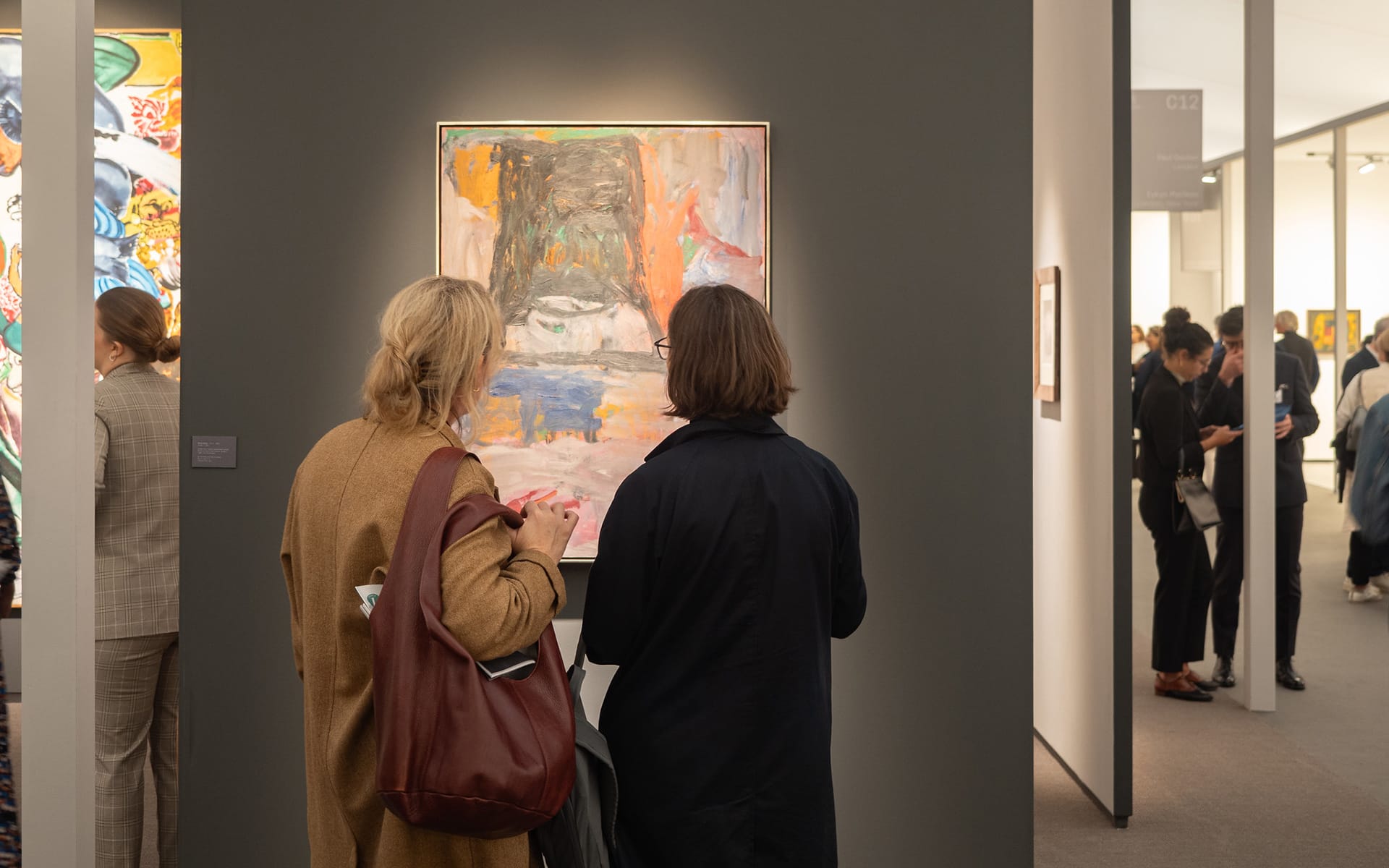 Two people viewing abstract art in a gallery setting with other visitors in the background.