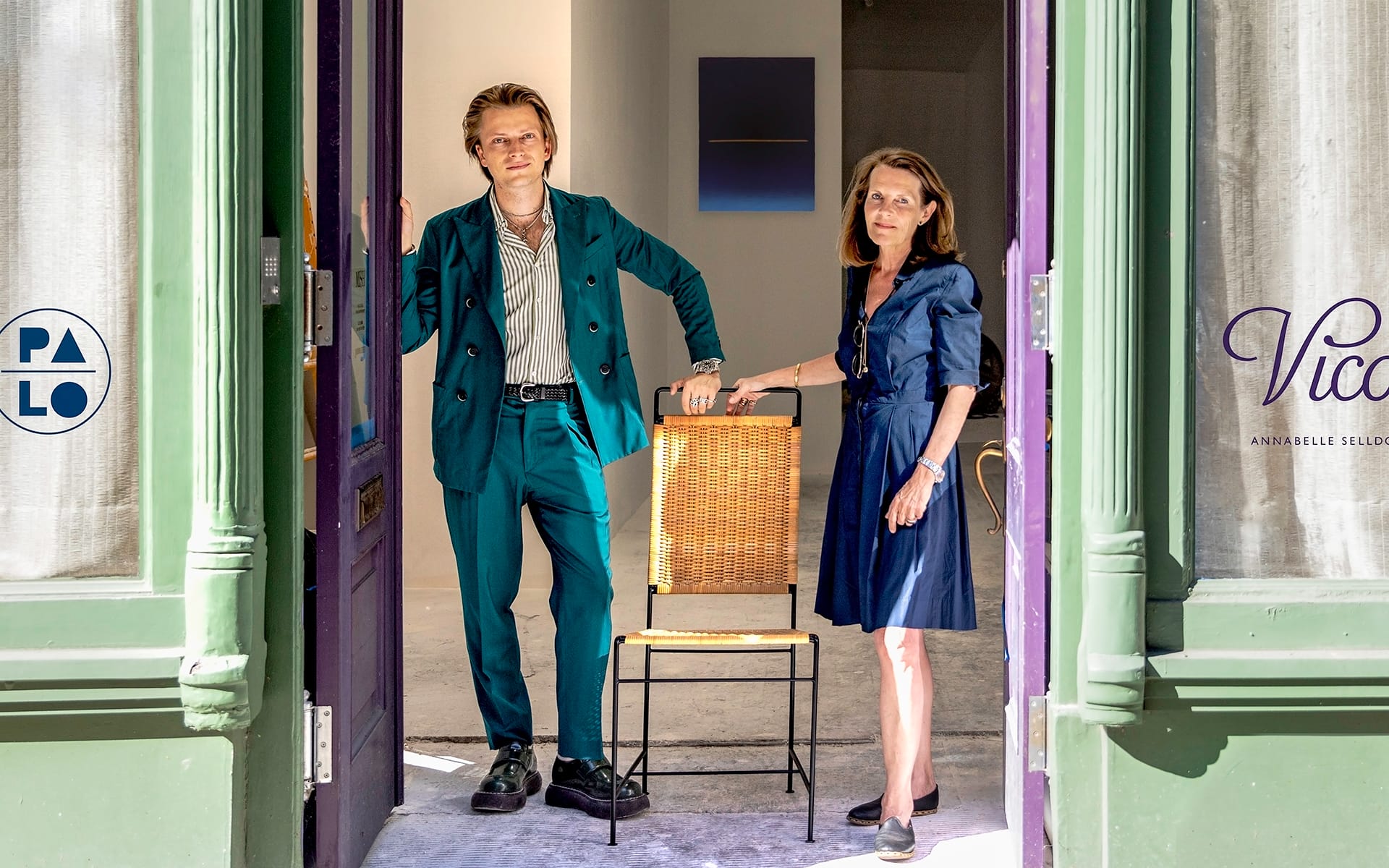 Two people standing at an open doorway, one in a green suit, the other in a blue dress, with a chair between them.