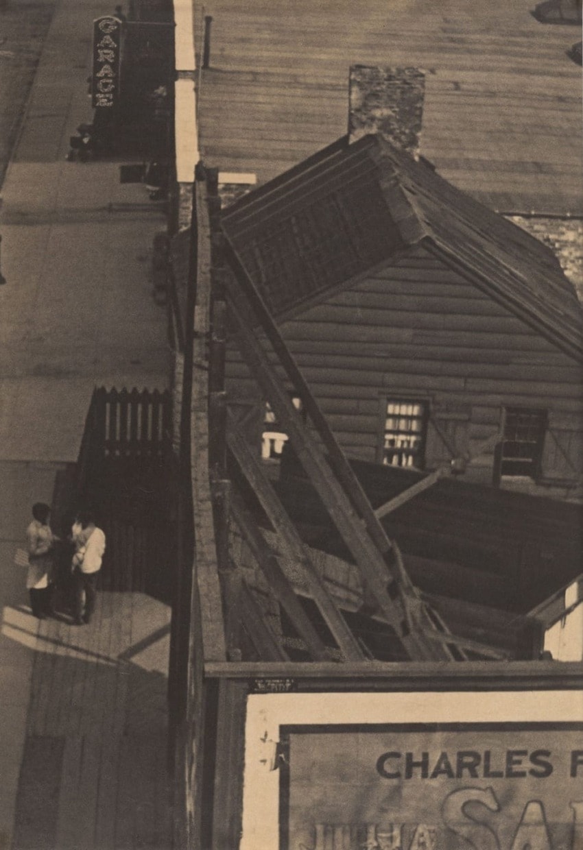Aerial view of people standing on a sidewalk next to a wooden building with a slanted roof and visible street signs.