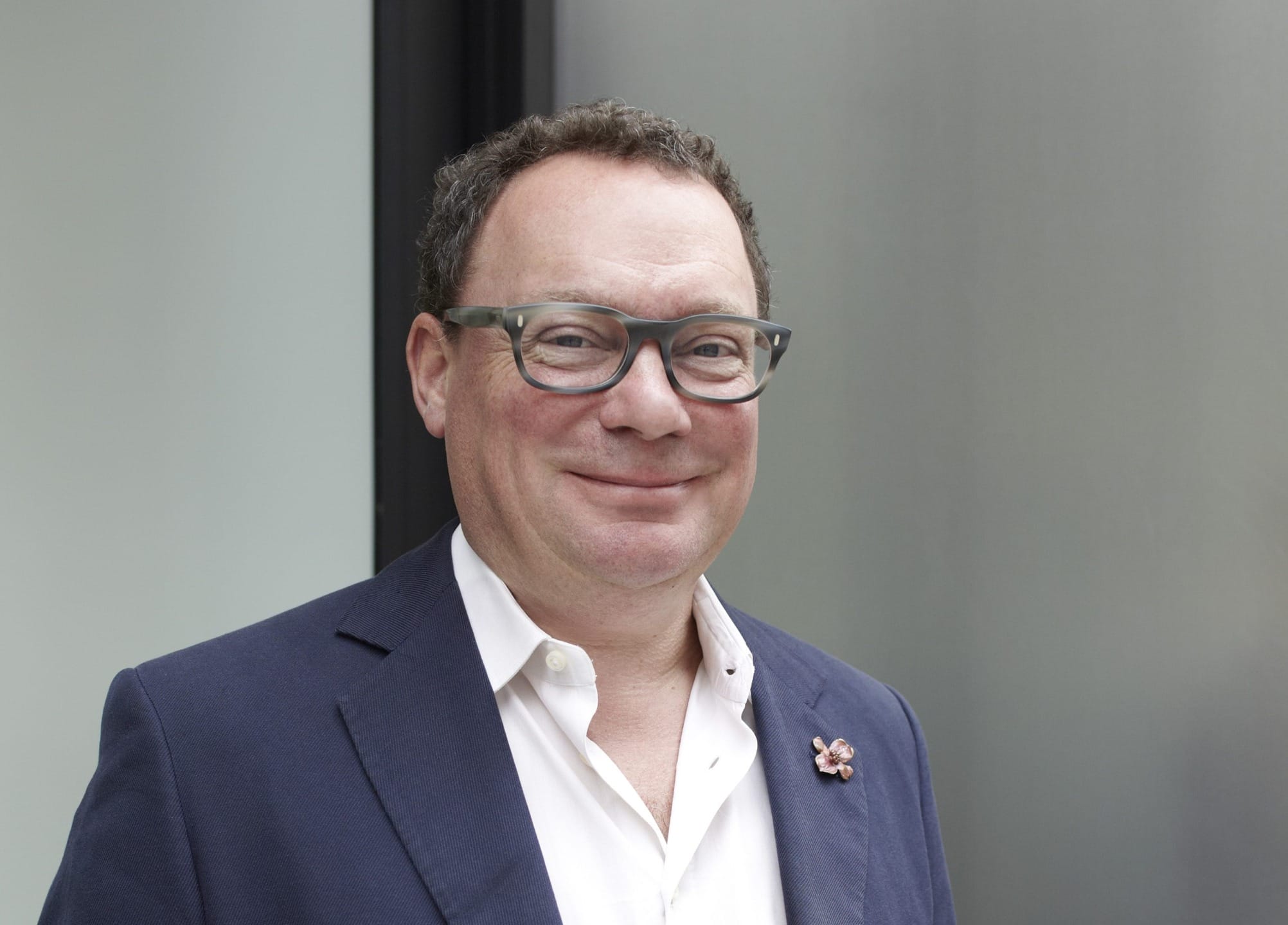 Man in glasses smiling, wearing a white shirt and dark jacket, standing in front of a neutral background.