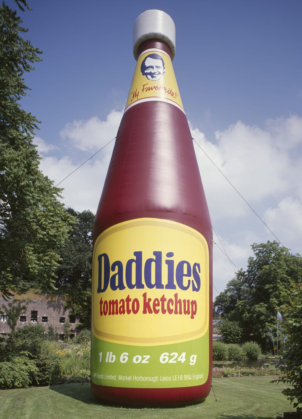 Giant inflated Daddies tomato ketchup bottle display outdoors with blue sky and trees in the background.
