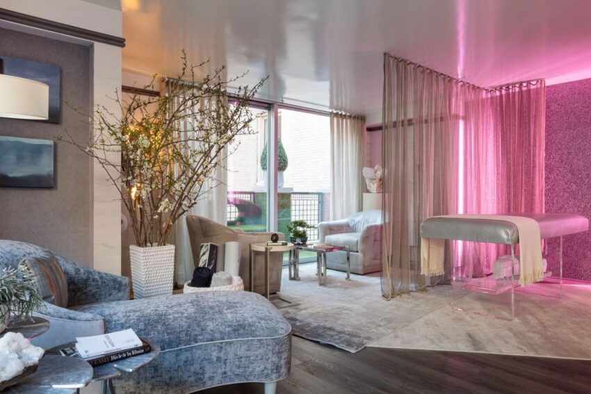 Modern spa room with lounge chairs, massage table, pink lighting, and large window with view of outdoor greenery.