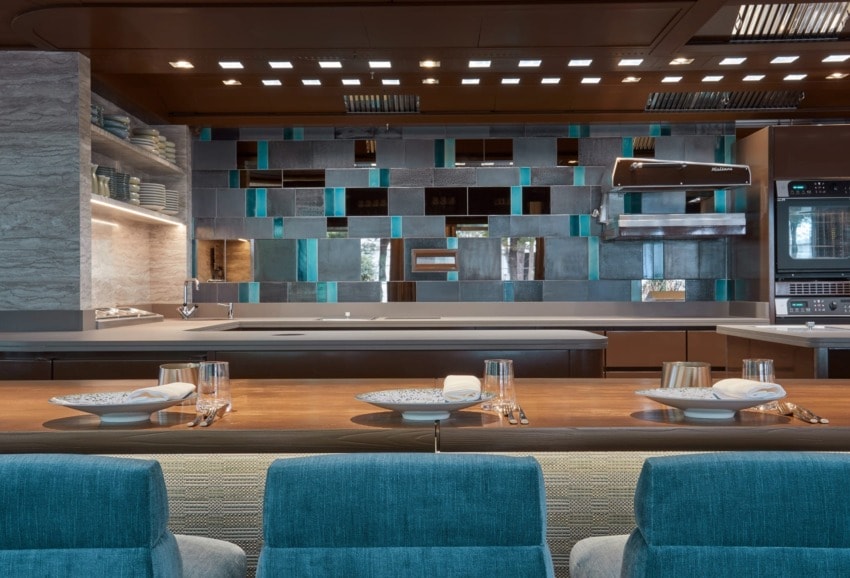 Modern kitchen with stylish backsplash, wooden counter, set dining space, blue chairs, and ambient lighting.