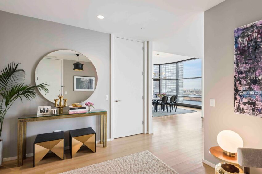 Modern apartment interior with decorative mirror, plants, and view into a dining area with large windows overlooking the city.