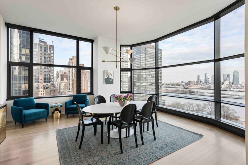 Modern dining room with large windows offering a cityscape view, a round table, black chairs, and teal upholstered armchairs.