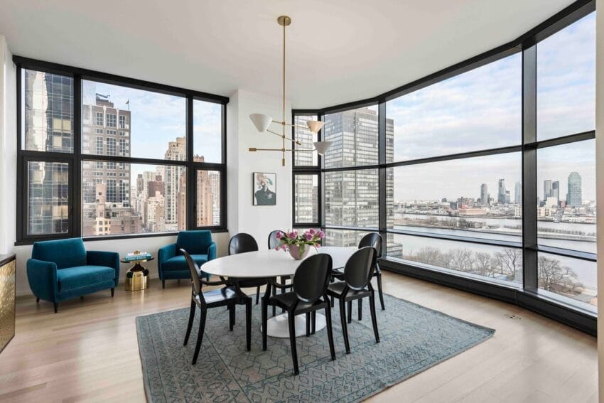 Modern dining room with large windows offering a cityscape view, a round table, black chairs, and teal upholstered armchairs.