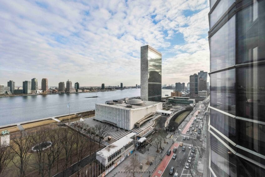 View of a cityscape featuring tall buildings, a riverfront, cloudy sky, and busy roads with cars and tree-lined sidewalks.