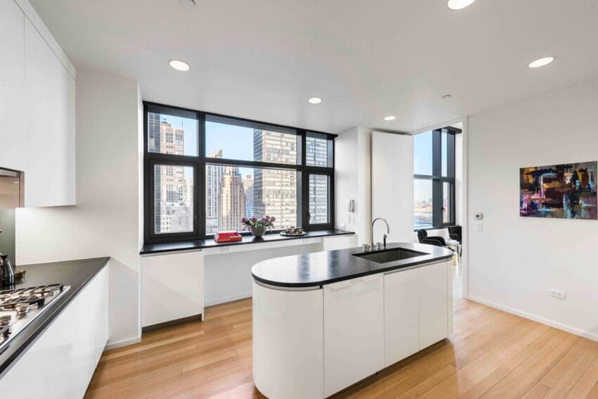 Modern kitchen with white cabinets, black countertops, island sink, and large windows overlooking cityscape.