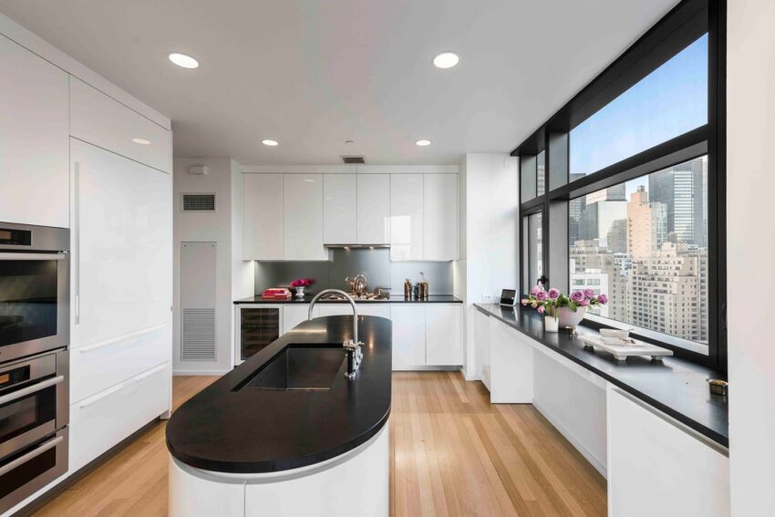 Modern kitchen with large window, city view, white cabinets, black countertops, island, built-in ovens, hardwood floors.