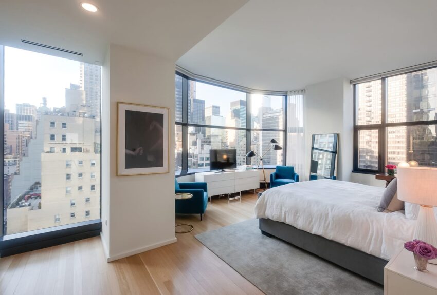 Spacious modern bedroom with large windows showcasing a city skyline, featuring a cozy bed and stylish armchairs.