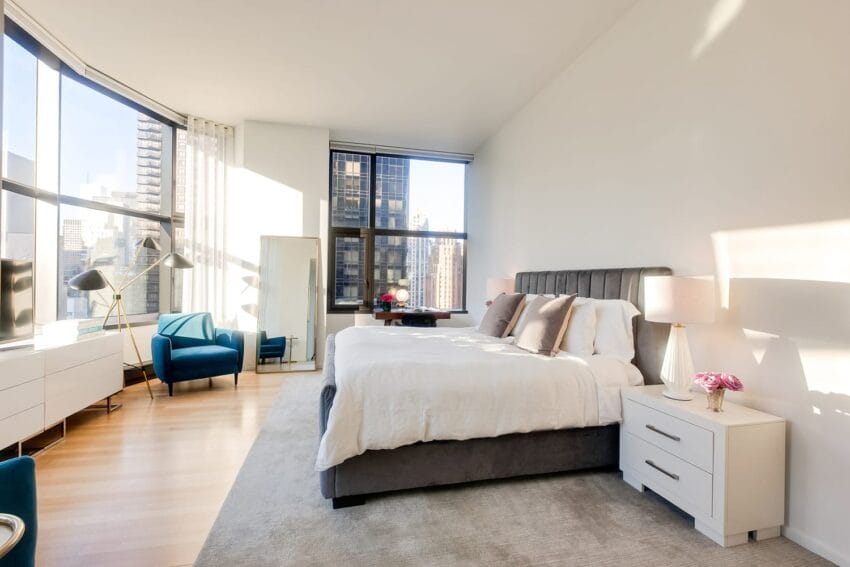 Spacious modern bedroom with large windows, city view, cozy bed, blue chairs, and stylish lamps.